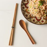 A pair of Teak Wood Chopsticks placed next to a spoon on a white surface, with a patterned bowl of rice and vegetables in the background.